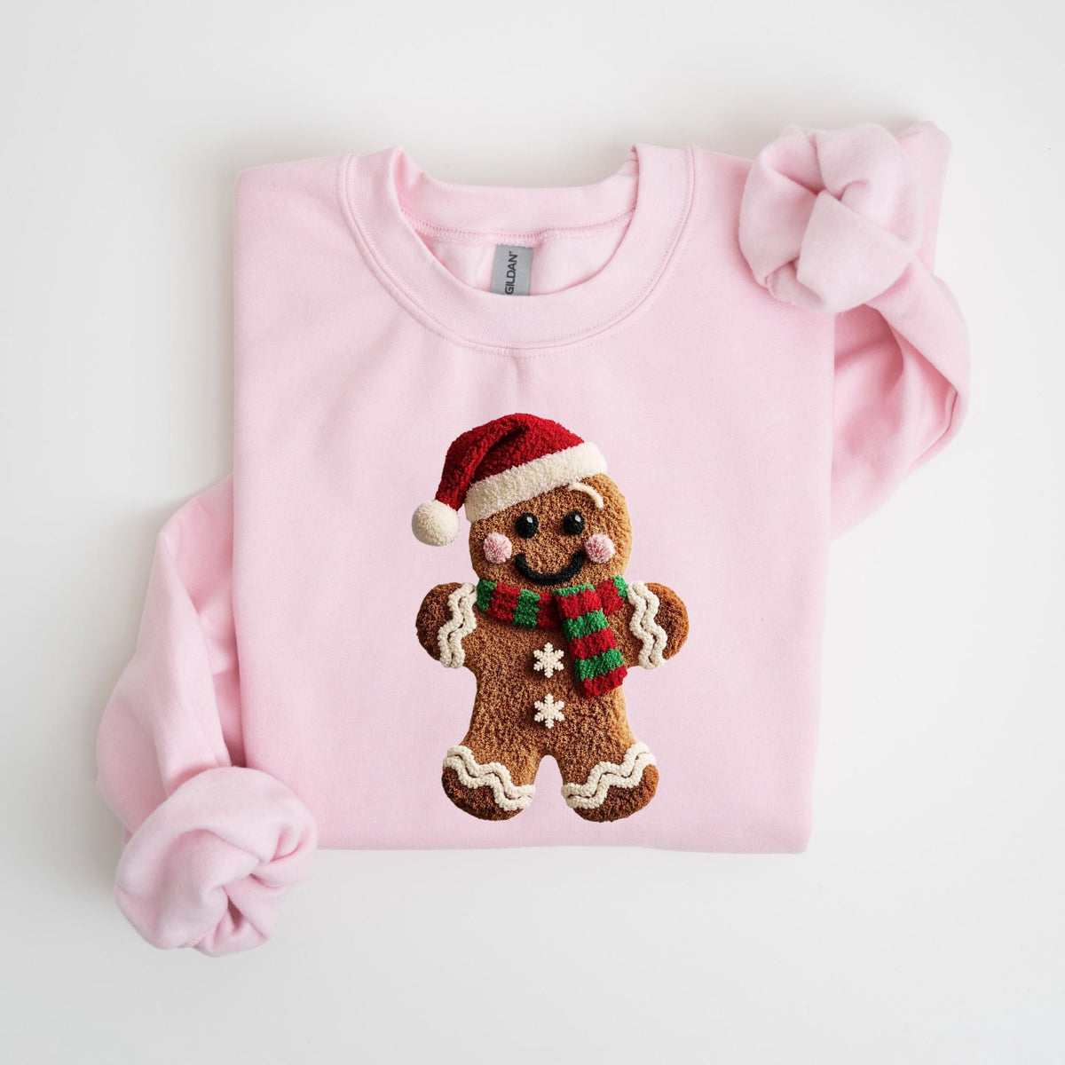 May include: A light pink sweatshirt featuring a gingerbread man design. The gingerbread man wears a red and white Santa hat and a red and green striped scarf. The gingerbread man has a smiling face and two white snowflake details.