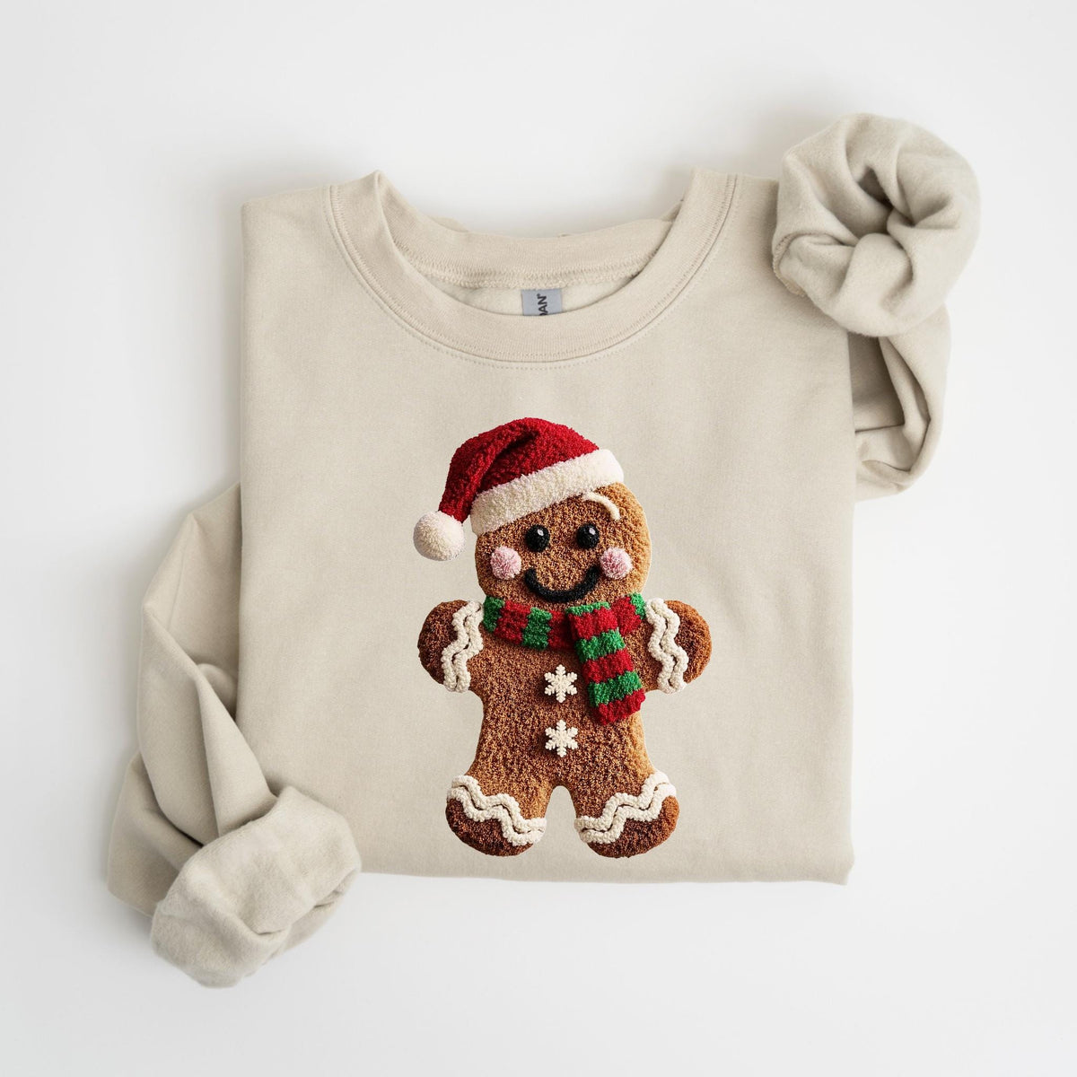 May include: A beige sweatshirt featuring a gingerbread man design. The gingerbread man wears a red Santa hat and a red and green scarf. The design includes white frosting details and two white snowflakes. The sweatshirt's sleeves are folded.