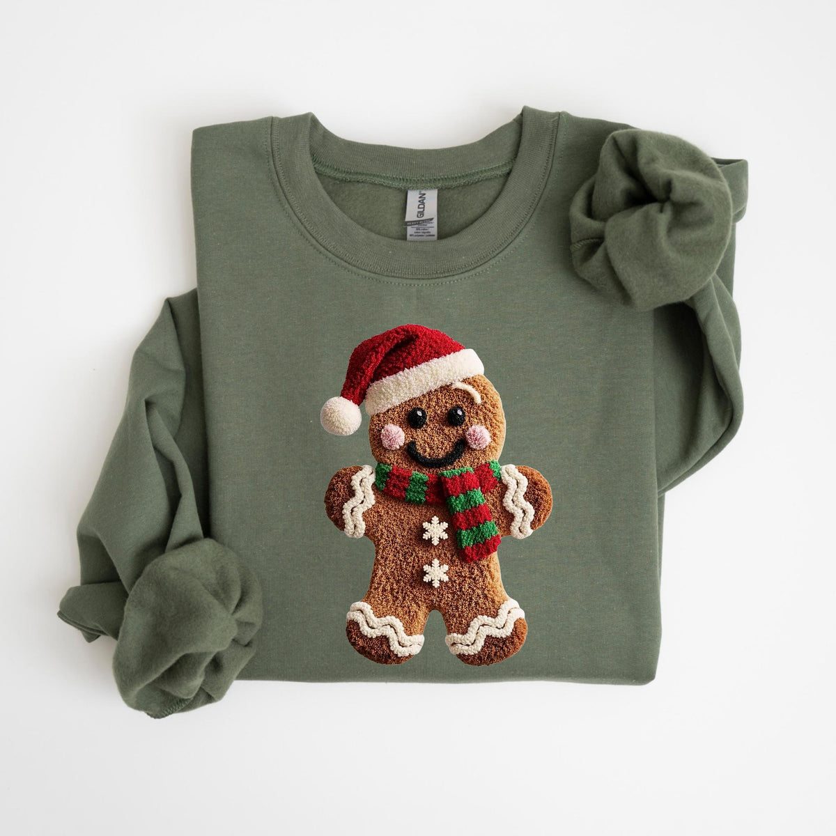 May include: An olive green sweatshirt featuring a gingerbread man design. The gingerbread man wears a red Santa hat and a red and green striped scarf. The gingerbread man has a smiling face and two white snowflakes.