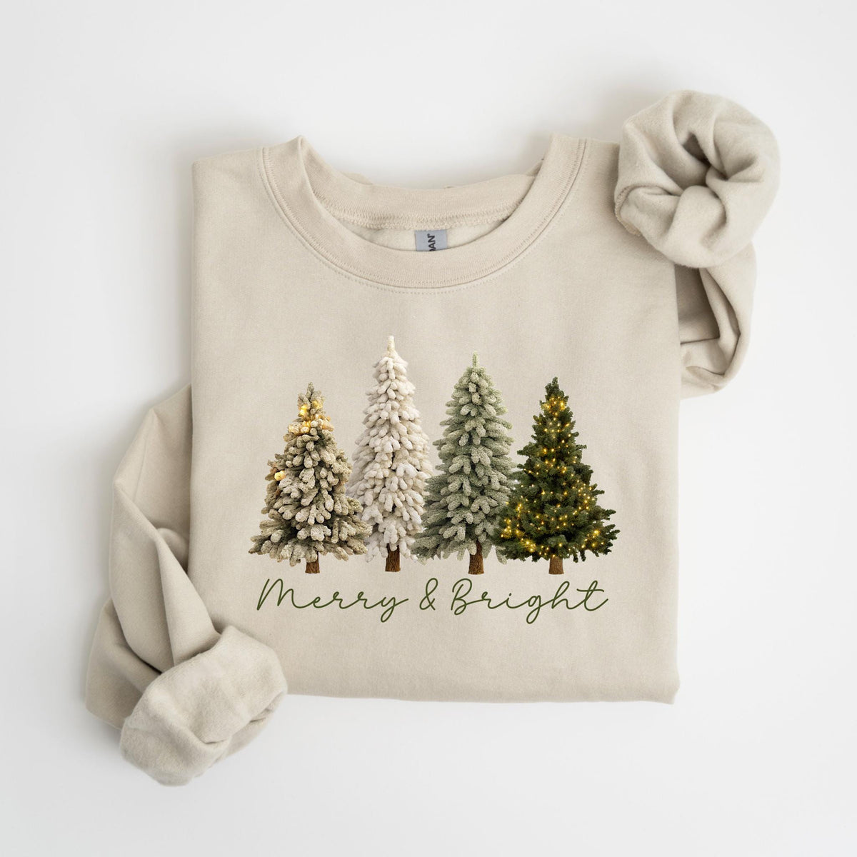 May include: A beige sweatshirt featuring four illustrated Christmas trees in varying shades of green and white, with some adorned with tiny lights. The phrase "Merry & Bright" is written in a script font below the trees. A matching scrunchie is placed on the top right.