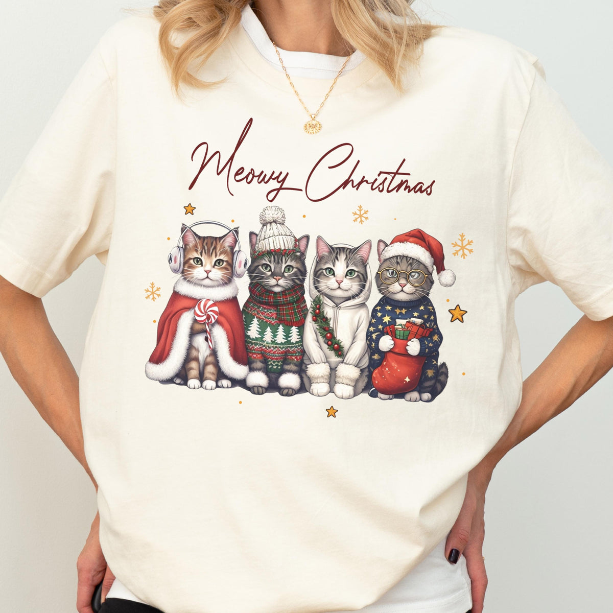 May include: A cream-colored t-shirt featuring a graphic of four cats dressed in winter holiday attire with the text Meowy Christmas. The cats wear accessories including earmuffs, a knit hat, a scarf, a hoodie, and a Santa hat with a stocking.