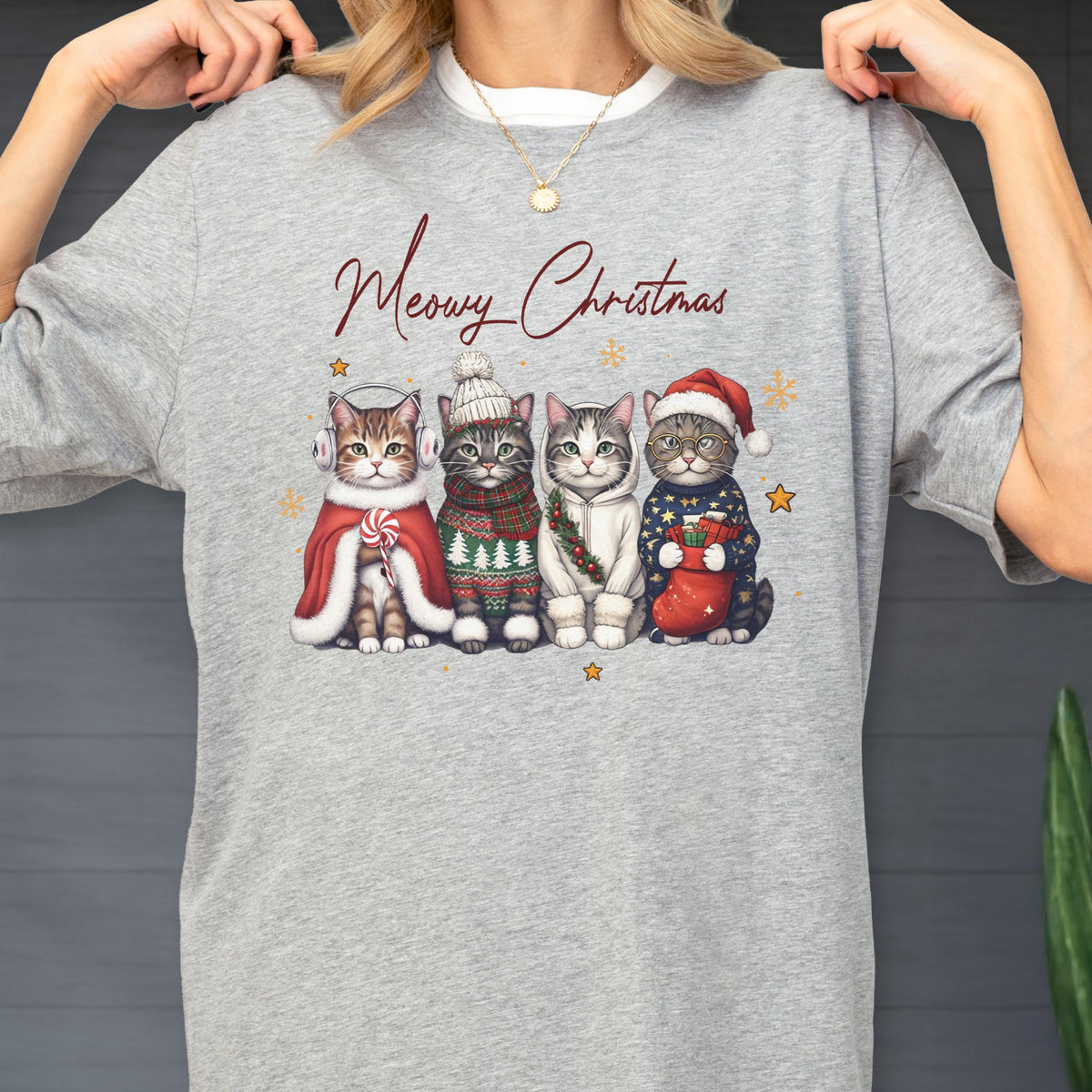 May include: A heather gray t-shirt featuring the text Meowy Christmas in red script above an illustration of four cats. The cats are wearing winter accessories including earmuffs, a knit hat, a scarf, a hoodie, and a Santa hat with a stocking.