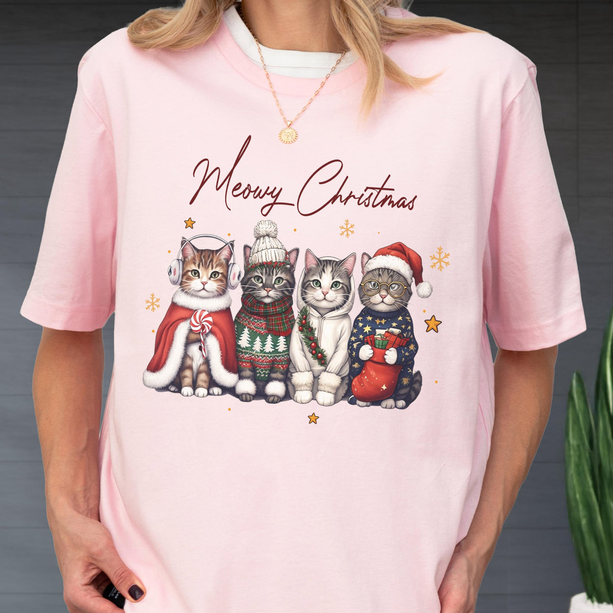 May include: A light pink t-shirt featuring the text Meowy Christmas in dark script above an illustration of four cats wearing winter accessories including earmuffs, a knit hat, a hoodie, and a Santa hat, accented by small gold stars and snowflakes.