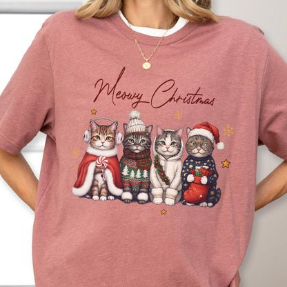 May include: A dusty rose t-shirt featuring the text Meowy Christmas in dark brown script above an illustration of four cats wearing winter accessories, including earmuffs, a knit hat, a hoodie, and a Santa hat, surrounded by small gold stars.