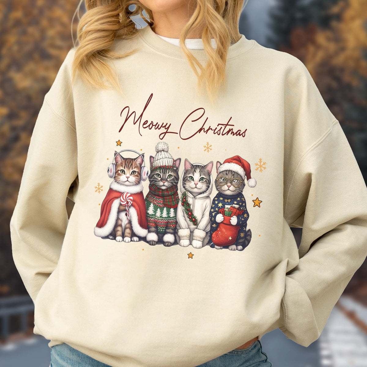 May include: A beige crewneck sweatshirt featuring the text Meowy Christmas in brown script above a graphic of four cats dressed in winter holiday attire, including earmuffs, a pom-pom beanie, a festive sweater, and a Santa hat with a stocking.