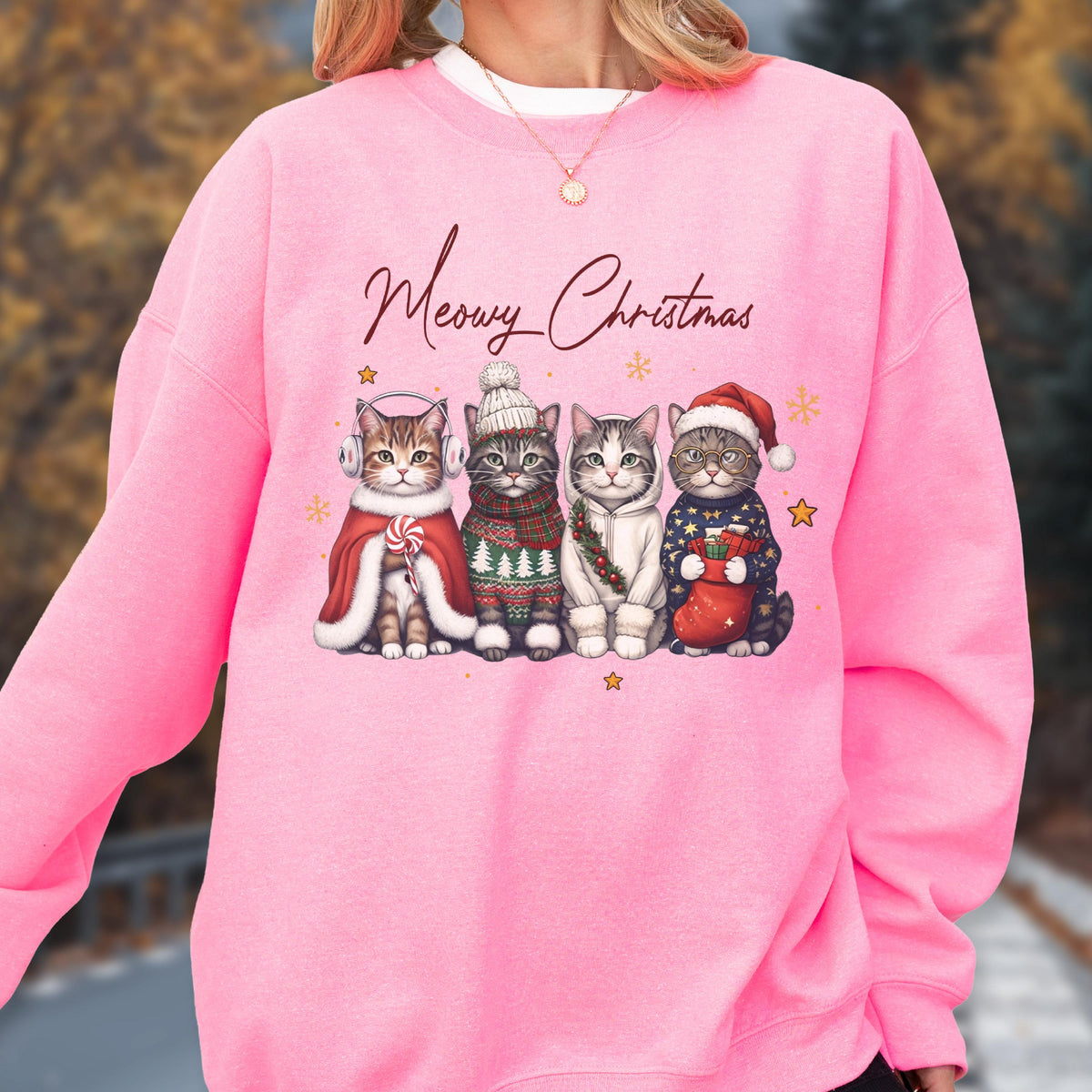 May include: A bright pink crewneck sweatshirt featuring a graphic of four cats dressed in winter attire with the text Meowy Christmas. The cats wear accessories including earmuffs, a knit hat, a scarf, and a Santa hat, surrounded by small gold stars.