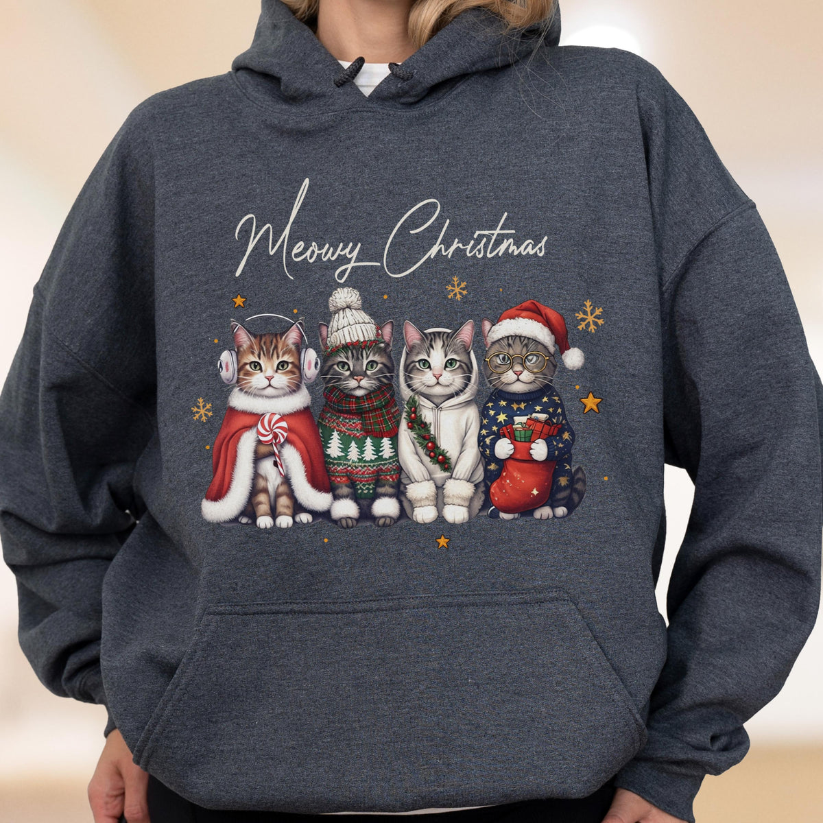 May include: A dark heather gray hooded sweatshirt featuring a graphic of four cats dressed in winter attire with the text Meowy Christmas. The cats wear accessories including earmuffs, a knit hat, a scarf, and a Santa hat, surrounded by stars.
