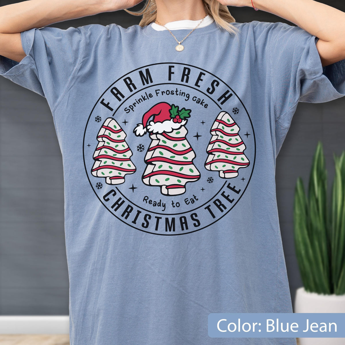 May include: A blue jean colored t-shirt featuring a circular graphic with the text FARM FRESH CHRISTMAS TREE, Sprinkle Frosting cake, and Ready to Eat. The design includes three white Christmas tree-shaped cakes with red stripes and green sprinkles.
