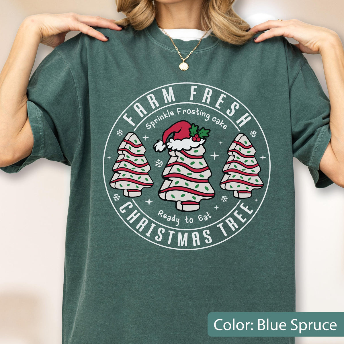 May include: A dark green t-shirt featuring a circular graphic with the text FARM FRESH CHRISTMAS TREE and Sprinkle Frosting Cake Ready to Eat. The design includes three white tree-shaped cakes with red stripes, green sprinkles, and a Santa hat.
