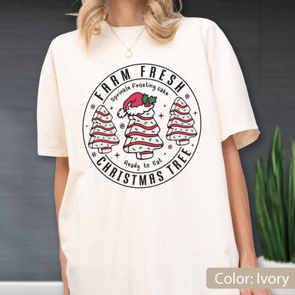 May include: An ivory short-sleeve t-shirt featuring a circular graphic with the text FARM FRESH CHRISTMAS TREE, Sprinkle Frosting cake, and Ready to Eat. The design includes three tree-shaped cakes with red stripes and green sprinkles.
