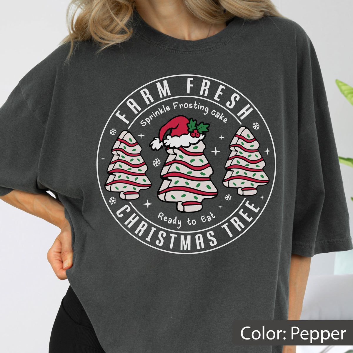 May include: A charcoal gray t-shirt featuring a circular graphic with the text FARM FRESH CHRISTMAS TREE. The design includes three tree-shaped cakes with white frosting, red stripes, and green sprinkles, with the center cake wearing a Santa hat.