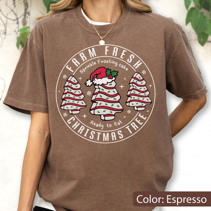 May include: An espresso-colored short-sleeve t-shirt featuring a circular graphic with the text FARM FRESH CHRISTMAS TREE, Sprinkle Frosting cake, and Ready to Eat. The design includes three white frosted tree-shaped cakes with red stripes and green sprinkles, with the center tree wearing a red Santa hat.