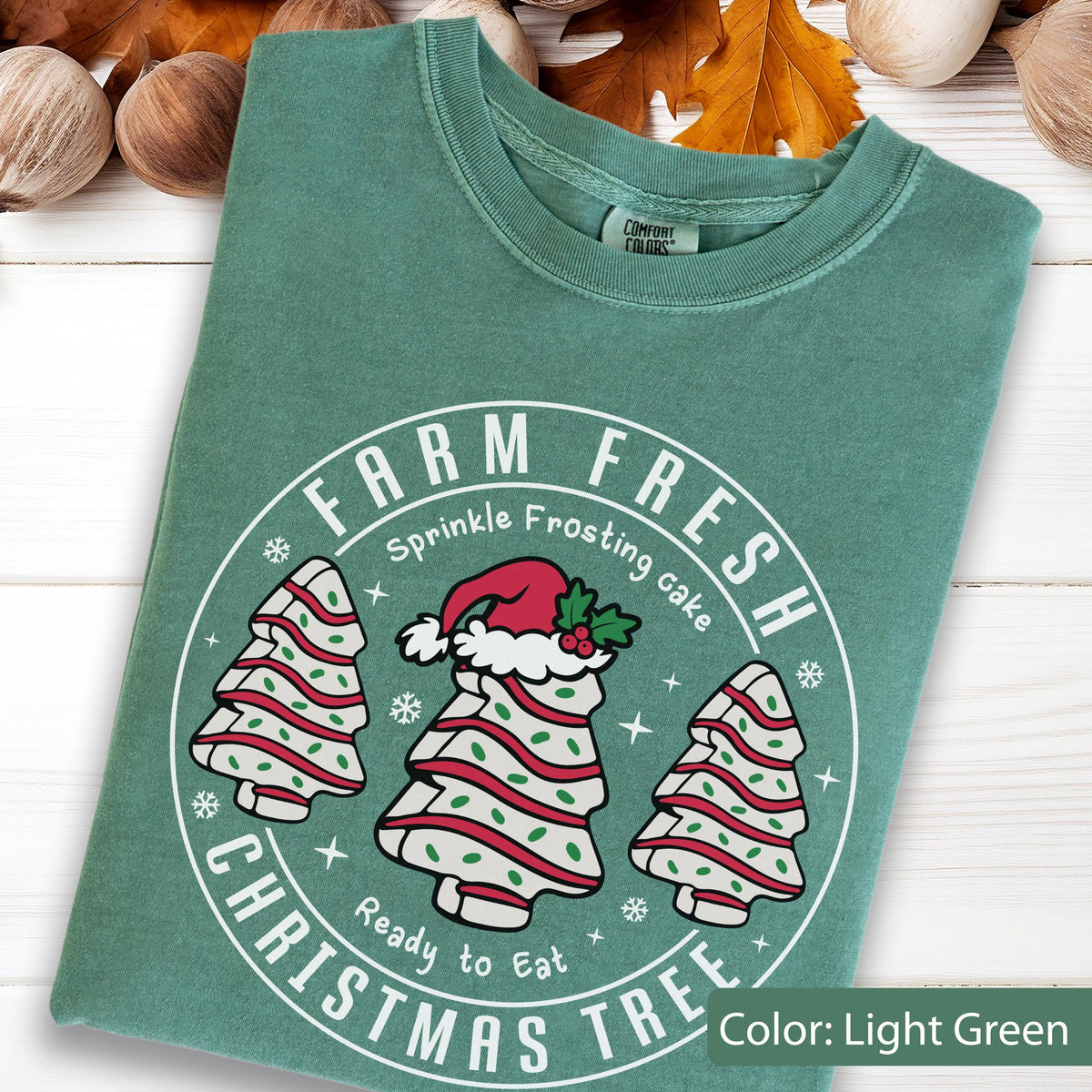 May include: A light green t-shirt featuring a white circular graphic with the text FARM FRESH CHRISTMAS TREE. The design includes three white tree-shaped cakes with red stripes and green sprinkles, with the center cake wearing a red Santa hat.