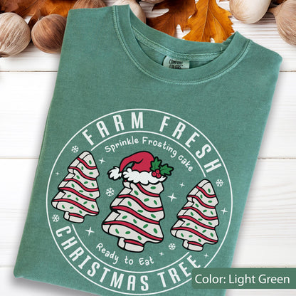 May include: A light green t-shirt featuring a white circular graphic with the text FARM FRESH CHRISTMAS TREE. The design includes three white tree-shaped cakes with red stripes and green sprinkles, with the center cake wearing a red Santa hat.