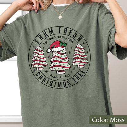 May include: A moss green t-shirt featuring a circular graphic with the text FARM FRESH, CHRISTMAS TREE, Sprinkle Frosting cake, and Ready to Eat. The design includes three white tree-shaped cakes with red stripes and green sprinkles, one wearing a Santa hat.