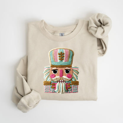 May include: Beige sweatshirt featuring a colorful embroidered nutcracker design. The nutcracker has a pink, blue, and gold color scheme. The sweatshirt has long sleeves and a crew neck.