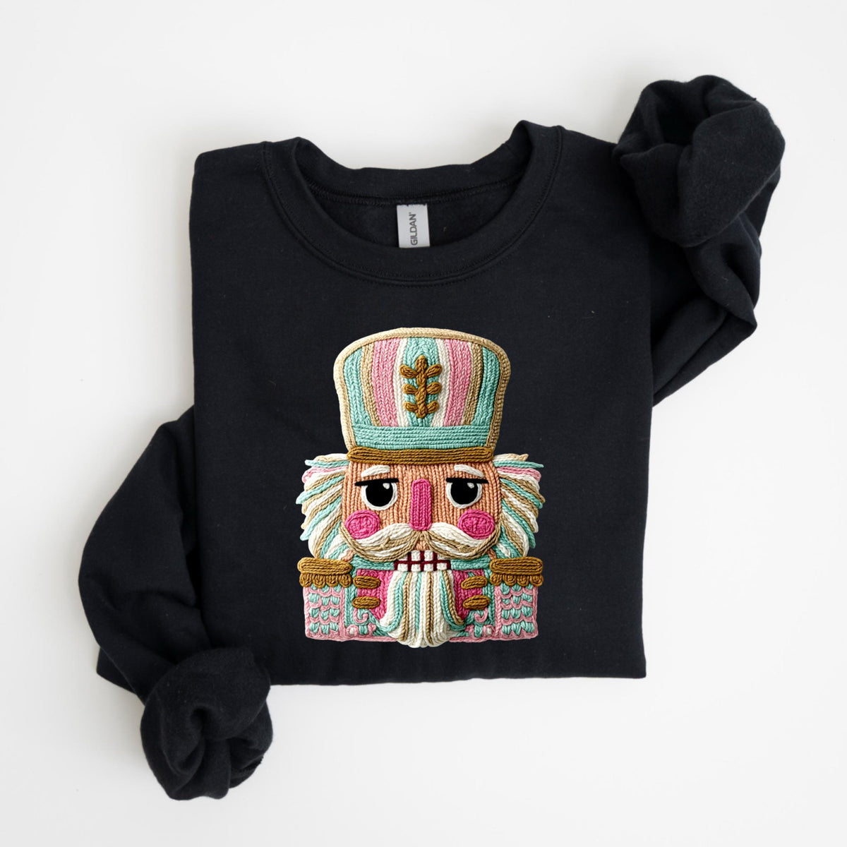 May include: Black crewneck sweatshirt featuring a colorful embroidered nutcracker design. The nutcracker has a pink face, a white mustache, and a detailed hat with pink, blue, and green stripes. The sweatshirt is folded neatly on a white surface.
