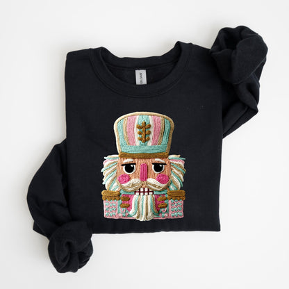May include: Black crewneck sweatshirt featuring a colorful embroidered nutcracker design. The nutcracker has a pink face, a white mustache, and a detailed hat with pink, blue, and green stripes. The sweatshirt is folded neatly on a white surface.