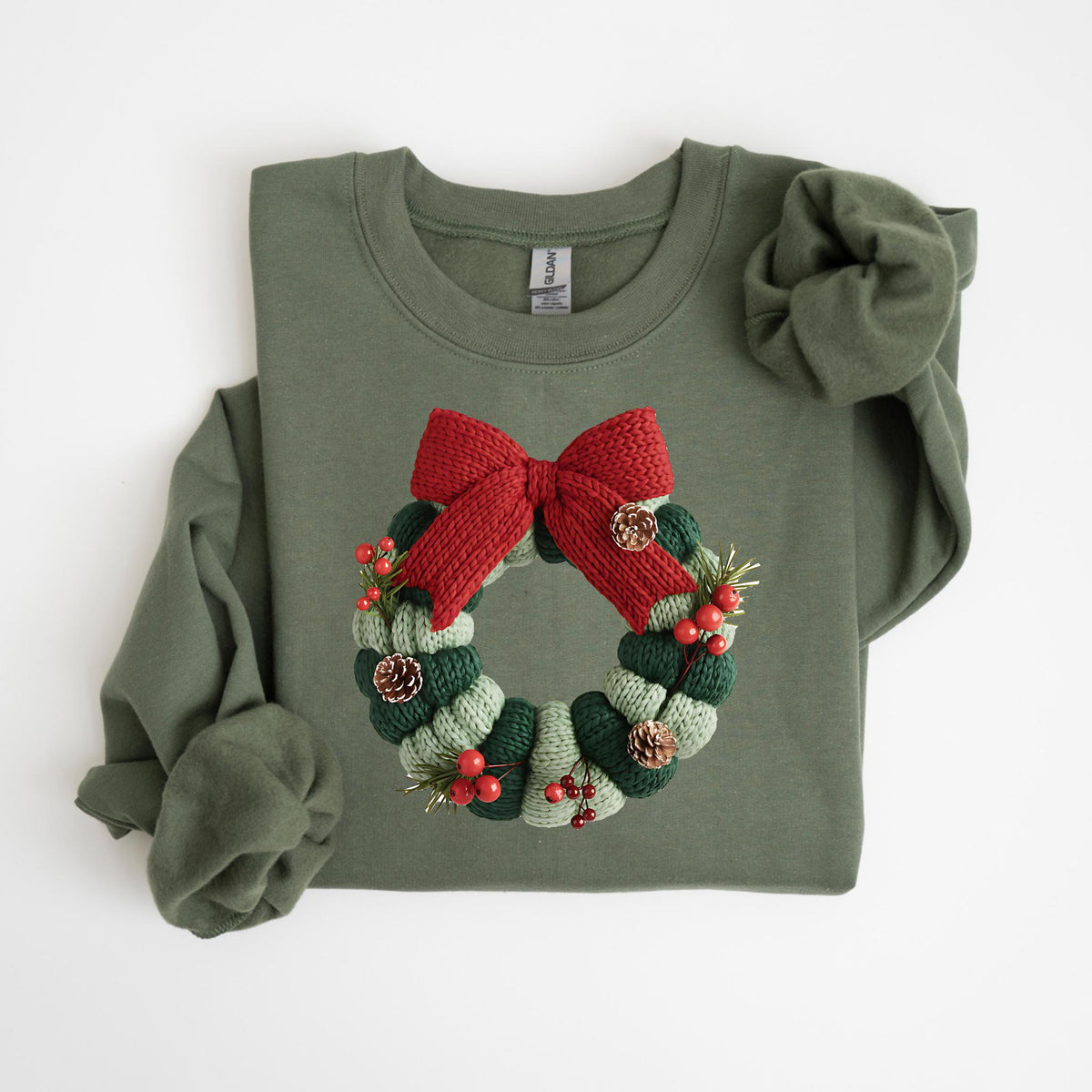 May include: Olive green sweatshirt featuring a knitted Christmas wreath design. The wreath is composed of green and white stripes, red berries, pinecones, and a red bow. A cozy, festive garment for the holiday season.