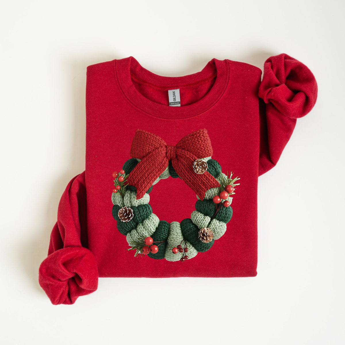 May include: A red sweatshirt featuring a knitted Christmas wreath design. The wreath is composed of green and light green sections, adorned with red berries, pinecones, and a large red bow at the top. The sleeves are folded.