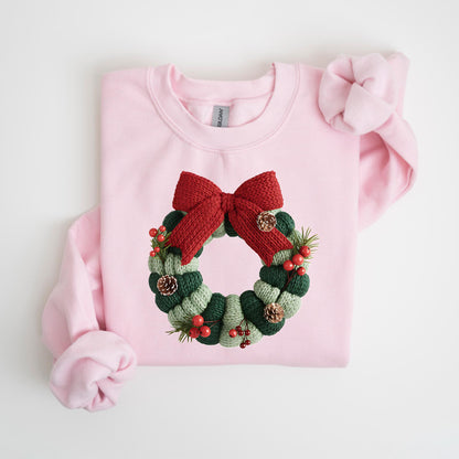 May include: A light pink sweatshirt featuring a knitted Christmas wreath design. The wreath is composed of green and light green sections, adorned with a red bow, pinecones, and red berries. The sweatshirt has long sleeves and a crew neck.