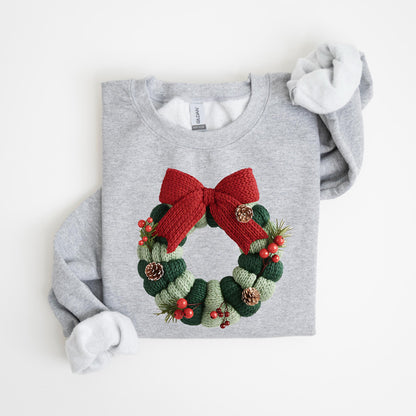 May include: A light gray sweatshirt featuring a knitted Christmas wreath design. The wreath is composed of green and white knitted sections, red berries, pinecones, and a large red bow. The sweatshirt has a crew neck and long sleeves.