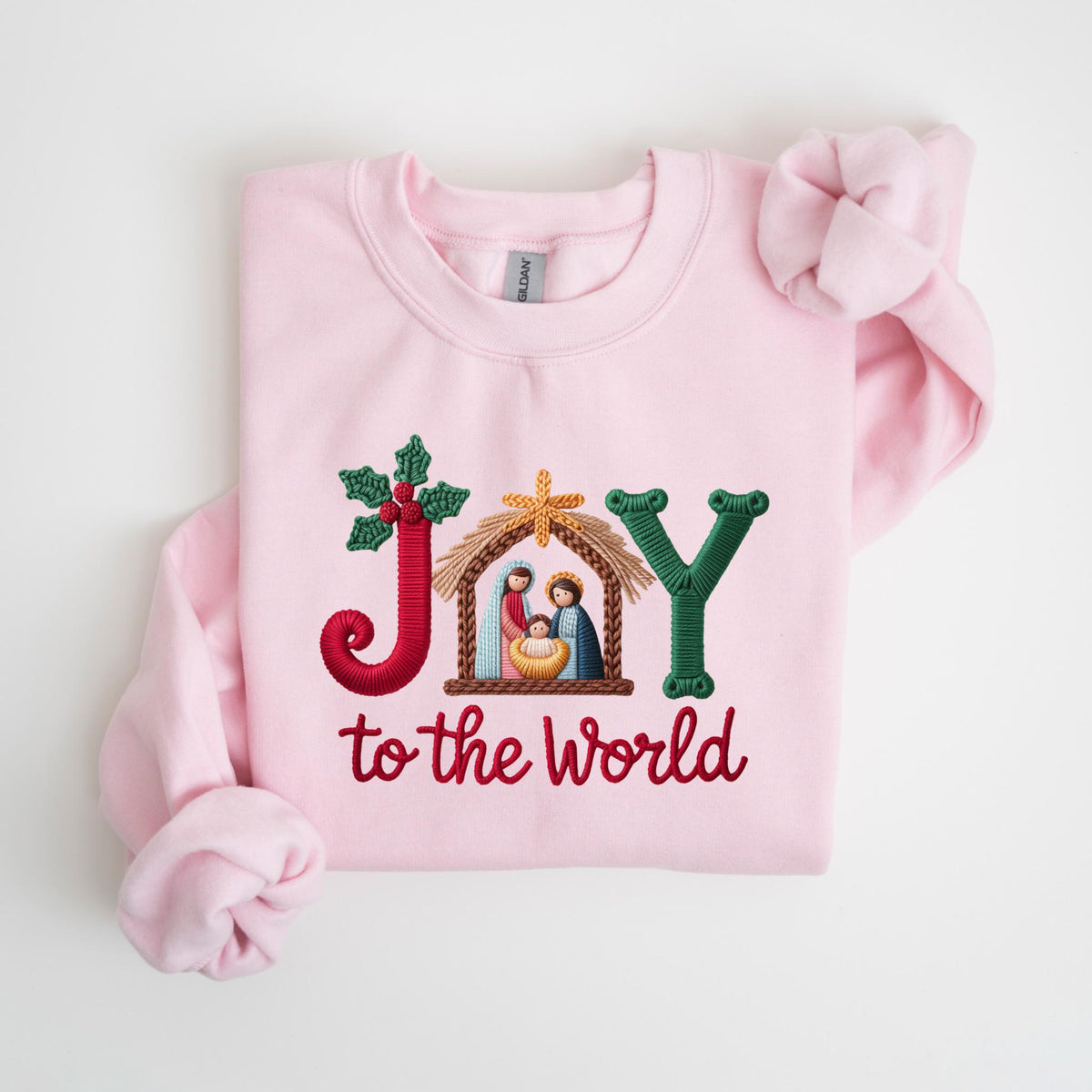 May include: A light pink sweatshirt with embroidered text and imagery. The word "JOY" is embroidered in red, green, and brown, with a nativity scene in the center. The phrase "to the world" is embroidered in red below the word "JOY".