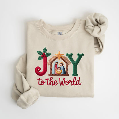 May include: Beige sweatshirt with embroidered text and design. The word "JOY" is embroidered in red, green, and gold, with a nativity scene in the center. The phrase "to the world" is embroidered in red below.
