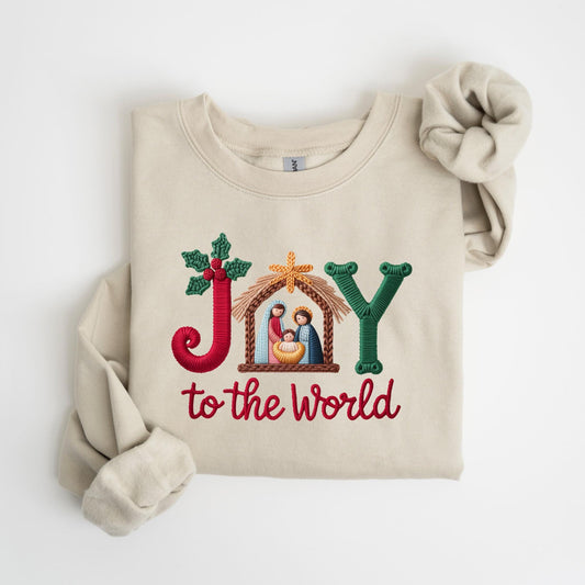May include: Beige sweatshirt with embroidered text and design. The word "JOY" is embroidered in red, green, and gold, with a nativity scene in the center. The phrase "to the world" is embroidered in red below.