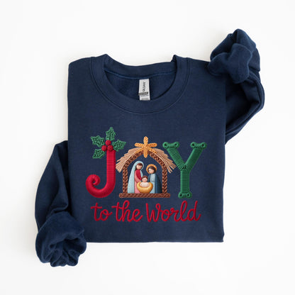May include: Navy blue sweatshirt with embroidered text and design. The design includes the word "JOY" with a nativity scene and the phrase "to the World" in red thread. The sweatshirt has long sleeves and a crew neck.