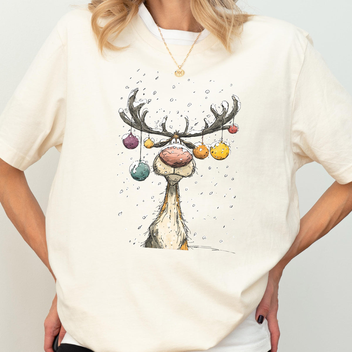 May include: Cream-colored t-shirt featuring a whimsical illustration of a reindeer with large antlers adorned with colorful ornaments. The reindeer has a large, pink nose and is set against a snowy background. The shirt has short sleeves.