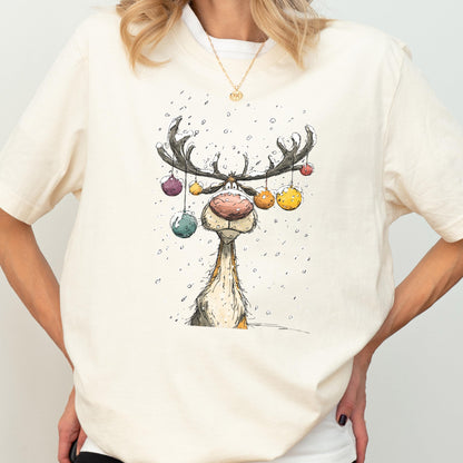 May include: Cream-colored t-shirt featuring a whimsical illustration of a reindeer with large antlers adorned with colorful ornaments. The reindeer has a large, pink nose and is set against a snowy background. The shirt has short sleeves.