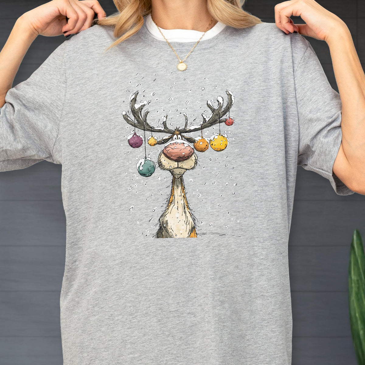 May include: A light gray t-shirt featuring a whimsical cartoon reindeer with colorful ornaments hanging from its antlers. The reindeer has a large pink nose and a long neck. The design is a festive, winter-themed graphic.