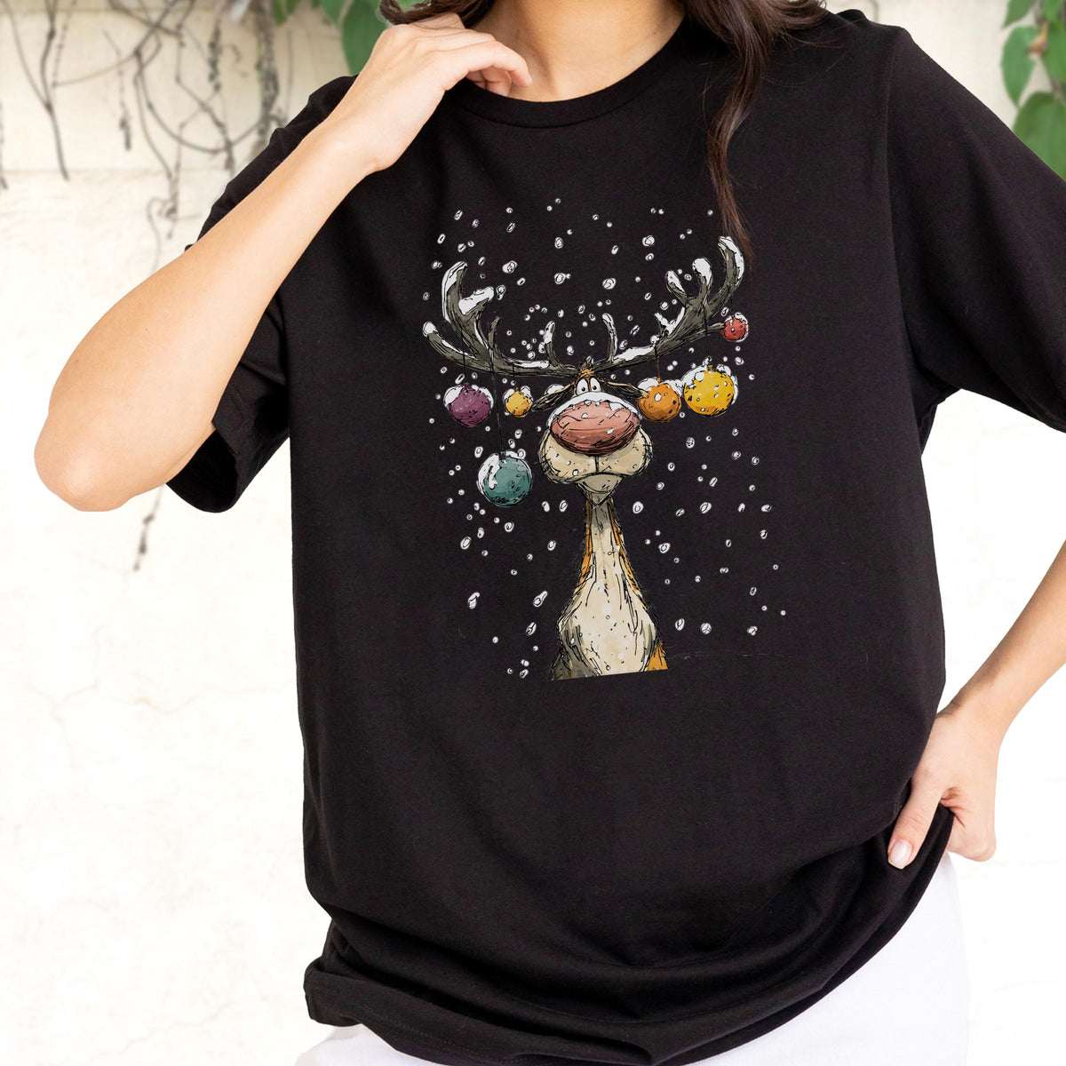 May include: Black t-shirt featuring a whimsical cartoon reindeer with large antlers adorned with colorful ornaments. The reindeer has a large red nose and is set against a snowy background, perfect for the holiday season.