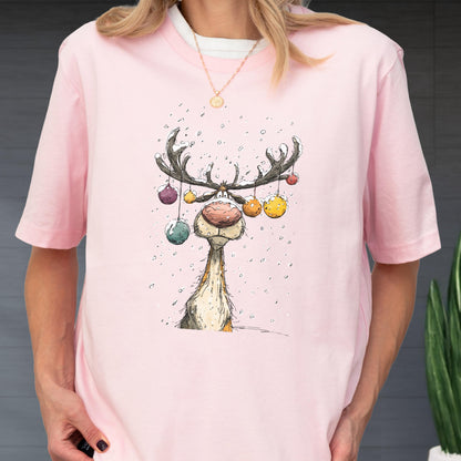 May include: A light pink t-shirt featuring a whimsical reindeer illustration. The reindeer has large antlers adorned with colorful ornaments and a prominent red nose. The design is set against a backdrop of falling snow, creating a festive holiday theme.