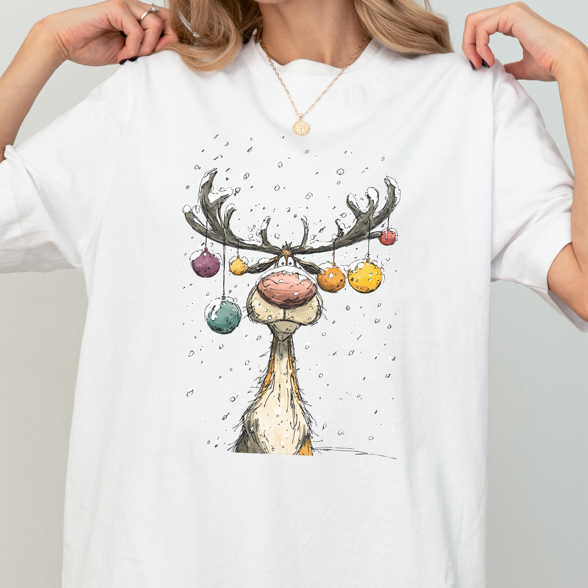May include: White t-shirt featuring a whimsical cartoon reindeer with large antlers adorned with colorful ornaments. The reindeer has a large, round nose and is set against a snowy background. A festive design for the holiday season.