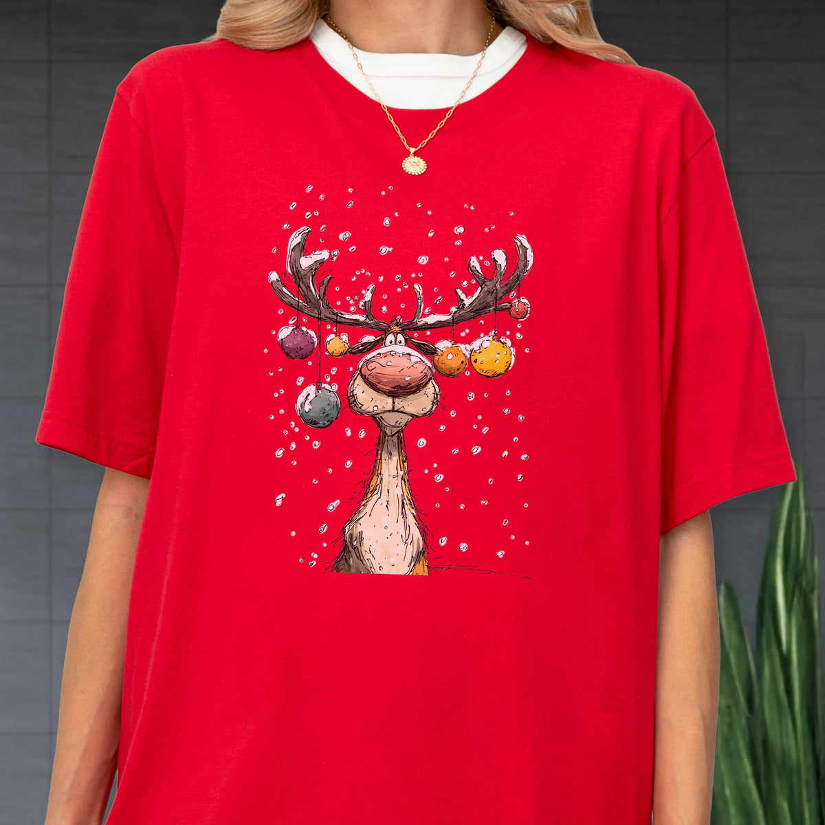 May include: Red t-shirt featuring a whimsical cartoon reindeer with large antlers and colorful ornaments. The reindeer has a large, red nose and is set against a snowy background. The shirt has a white crew neck.