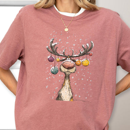 May include: A dusty rose t-shirt featuring a whimsical reindeer illustration. The reindeer has large antlers adorned with colorful ornaments. Snowflakes are falling around the reindeer. The shirt has a crew neck and short sleeves.
