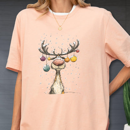 May include: A light pink t-shirt featuring a whimsical reindeer illustration. The reindeer has large antlers adorned with colorful ornaments and a large, pink nose. Snowflakes are falling around the reindeer. The shirt has a white crew neck.