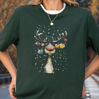 May include: Dark green t-shirt featuring a cartoon reindeer with colorful ornaments on its antlers and falling snow. The reindeer has a large red nose and a whimsical expression. The shirt has a white crew neck.