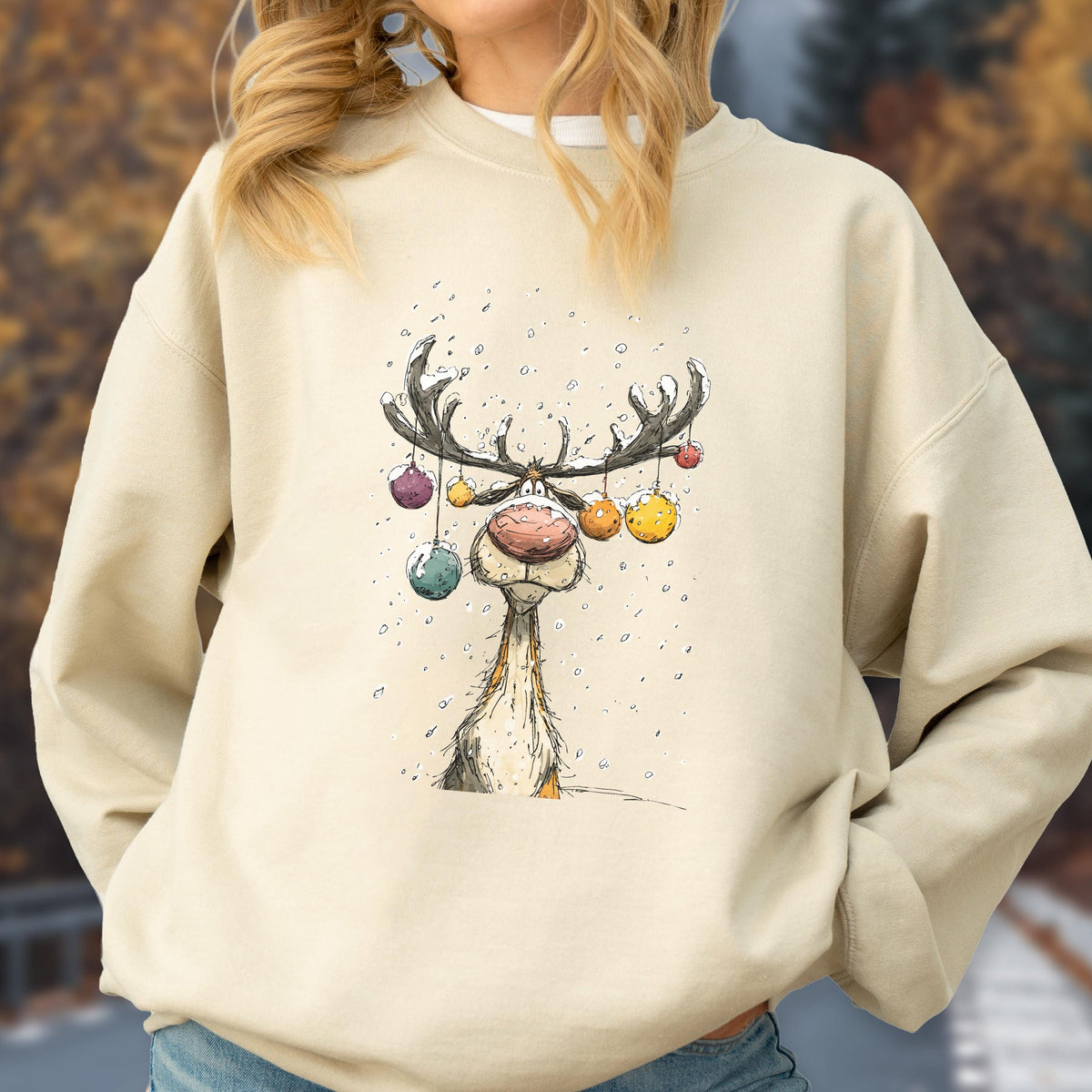 May include: A beige crewneck sweatshirt featuring a graphic illustration of a reindeer with large antlers decorated with colorful hanging ornaments. The reindeer has a prominent pink nose and is surrounded by small white dots representing snow.