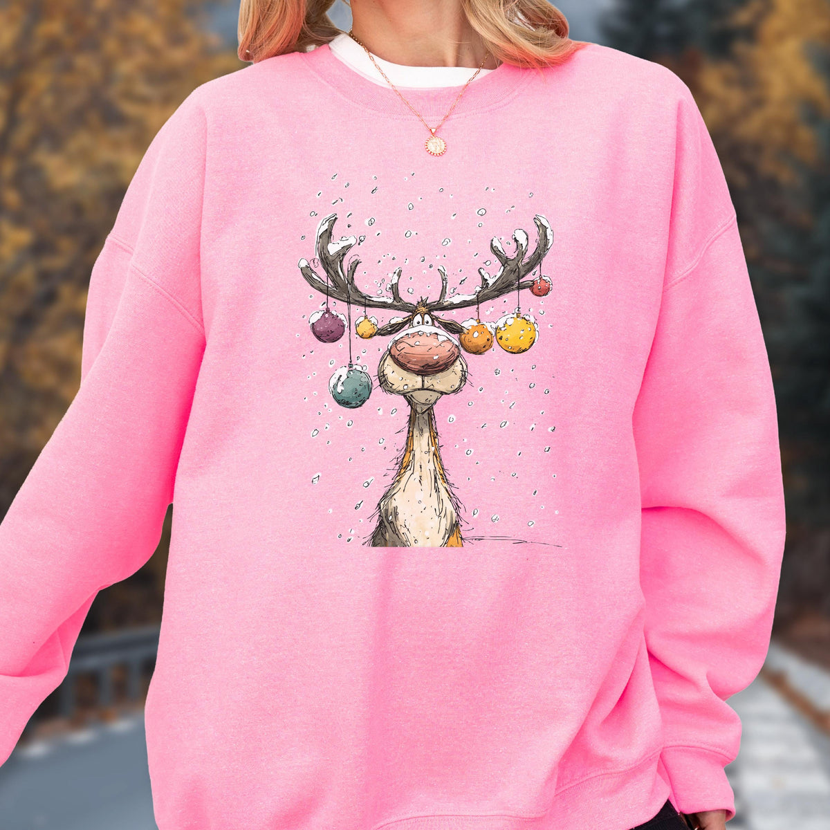 May include: A bright pink long-sleeve sweatshirt featuring a central graphic illustration of a reindeer with large antlers decorated with colorful hanging ornaments. The reindeer has a prominent nose and is surrounded by small white dots resembling snow.