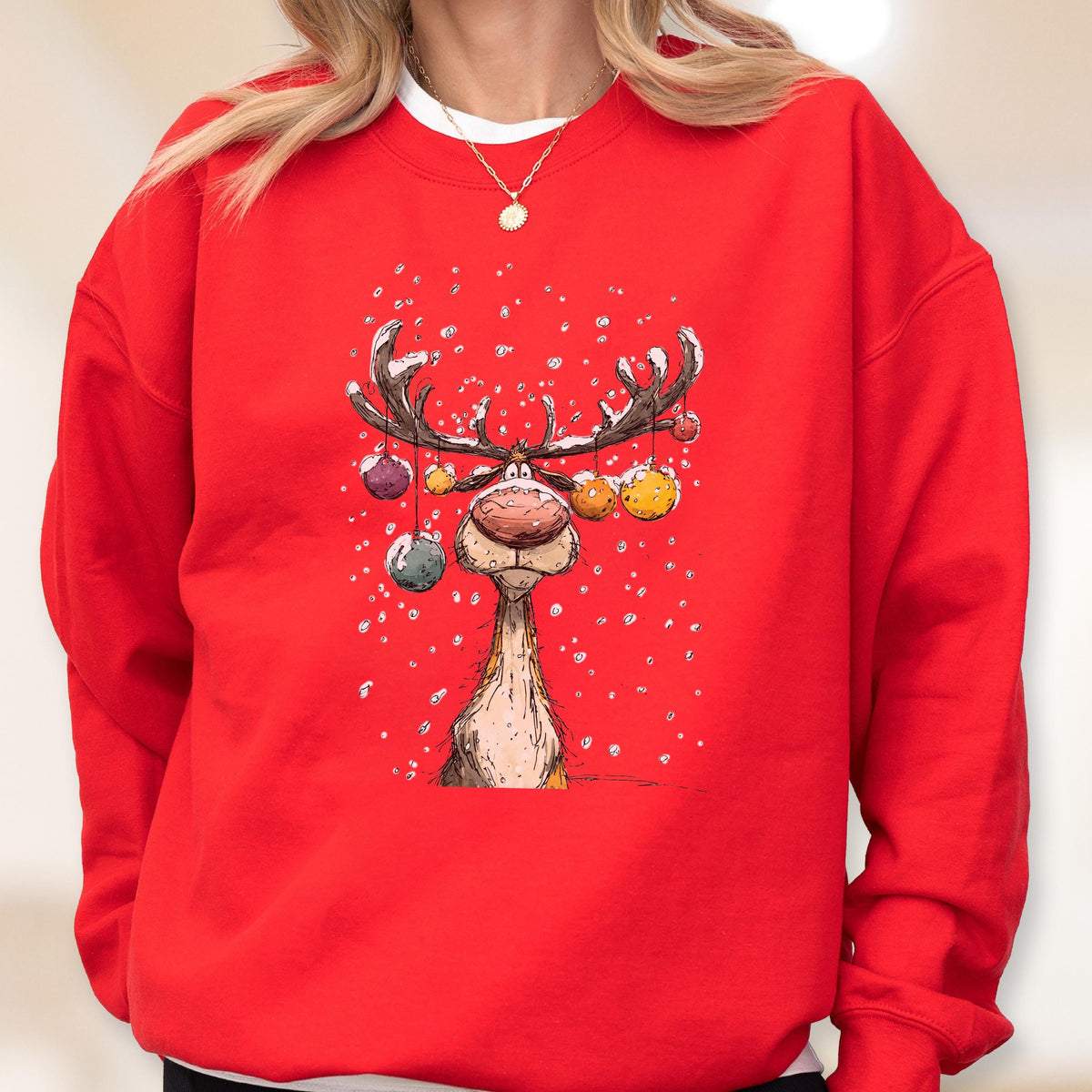 May include: A bright red crewneck sweatshirt featuring a graphic illustration of a cartoon reindeer with large antlers decorated with colorful hanging ornaments, set against a backdrop of white falling snow, with a white undershirt collar visible.