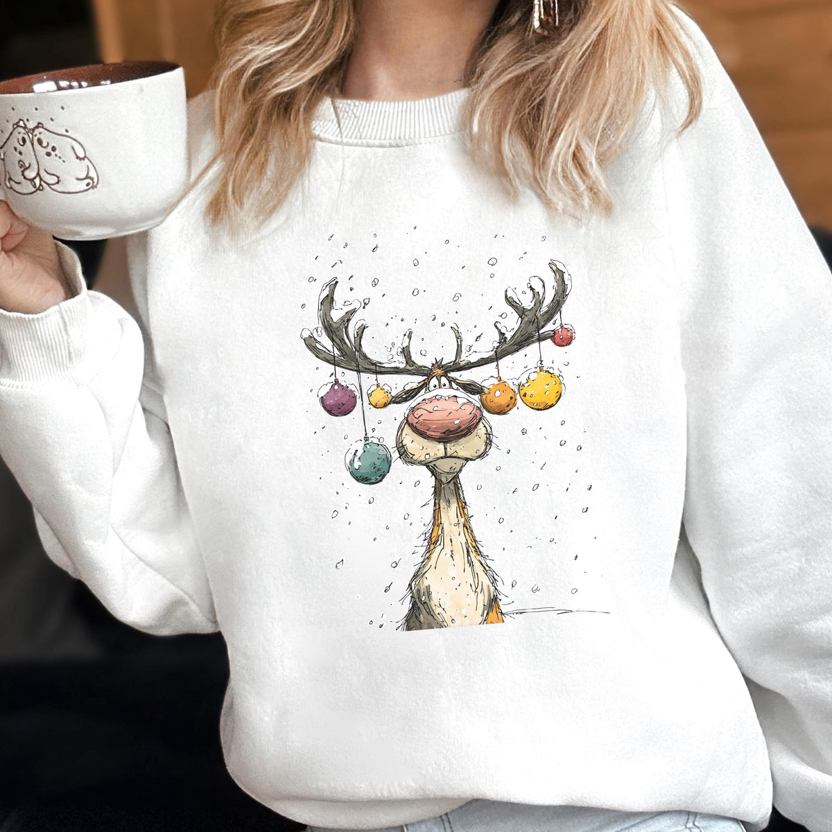 May include: A white crewneck sweatshirt featuring a graphic illustration of a reindeer with a large pink nose and antlers decorated with colorful hanging ornaments, set against a backdrop of falling snow, paired with a matching white ceramic mug.