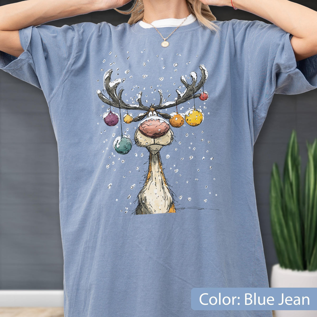 May include: A blue jean colored t-shirt featuring a graphic illustration of a reindeer with a large nose and antlers decorated with colorful hanging ornaments. The design includes white specks representing falling snow around the reindeer.