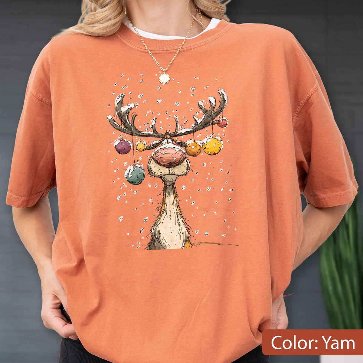 May include: A burnt orange crewneck t-shirt featuring a graphic illustration of a reindeer with large antlers decorated with colorful hanging ornaments. The reindeer has a prominent nose and is surrounded by white dots representing falling snow.