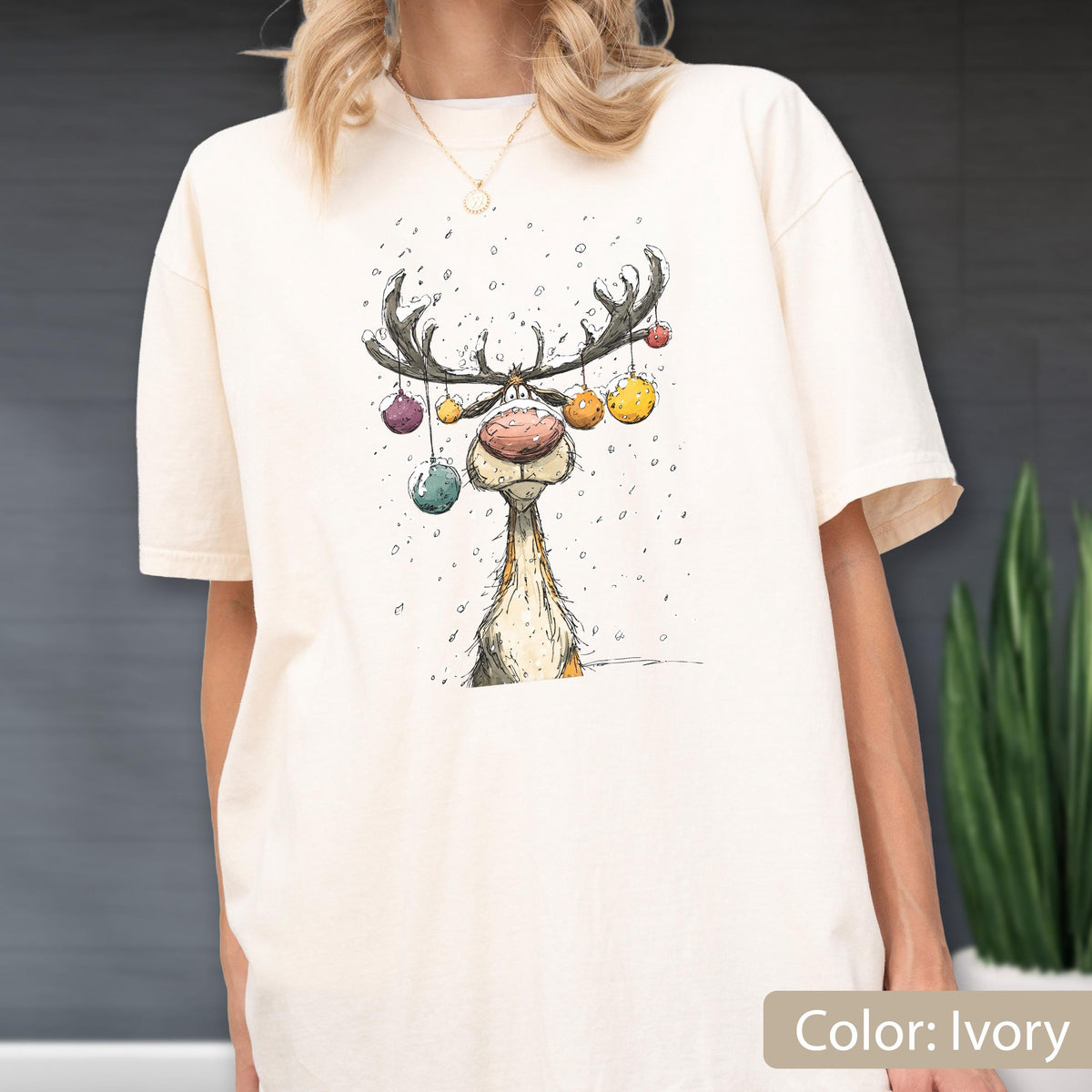 May include: An ivory short-sleeve t-shirt featuring a graphic illustration of a reindeer with a large pink nose. The reindeer has colorful Christmas ornaments hanging from its antlers, and small white dots representing falling snow.