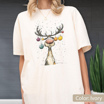 May include: An ivory short-sleeve t-shirt featuring a graphic illustration of a reindeer with a large pink nose. The reindeer has colorful Christmas ornaments hanging from its antlers, and small white dots representing falling snow.