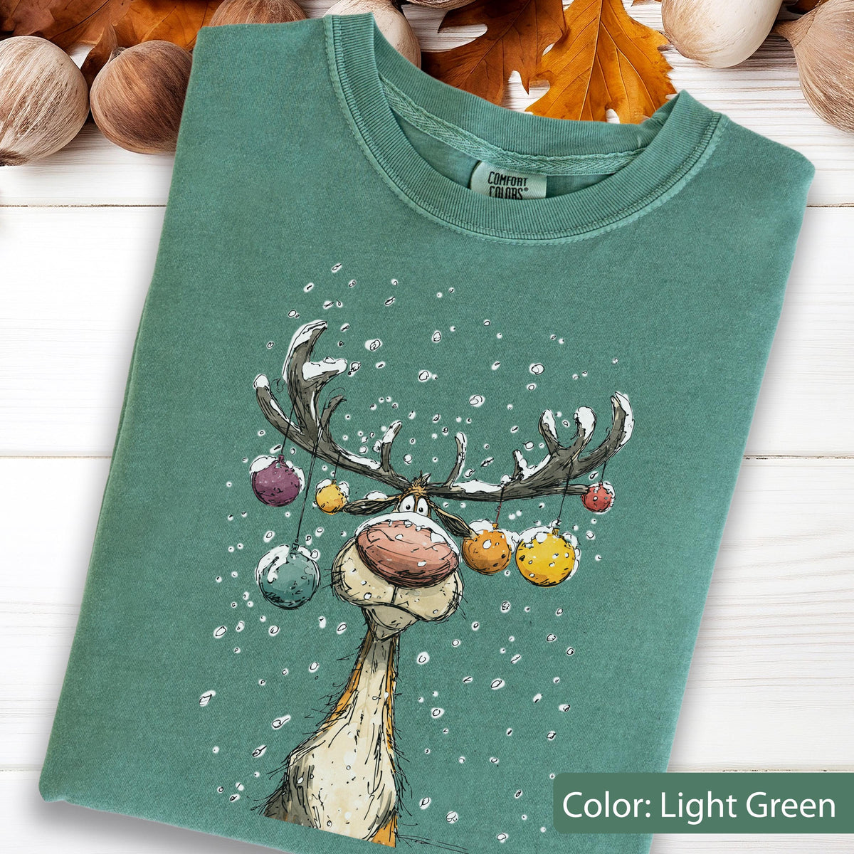 May include: A light green crewneck t-shirt featuring a graphic illustration of a reindeer with large antlers decorated with colorful hanging ornaments. The reindeer has a large nose and is surrounded by white snowflakes. The shirt has a Comfort Colors label.