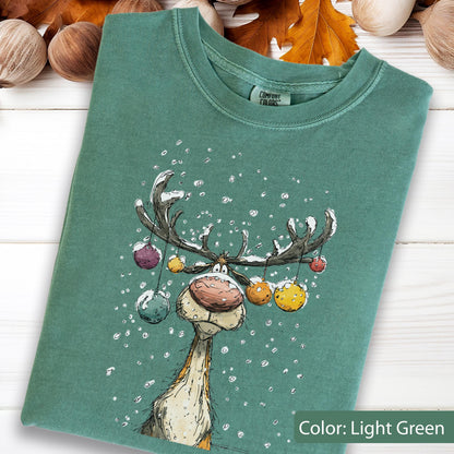 May include: A light green crewneck t-shirt featuring a graphic illustration of a reindeer with large antlers decorated with colorful hanging ornaments. The reindeer has a large nose and is surrounded by white snowflakes. The shirt has a Comfort Colors label.
